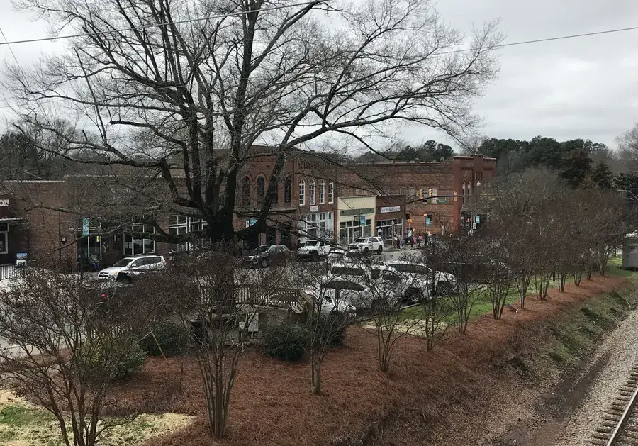 A row of brick shops in downtown Waxhaw seen across a planted strip, with the railroad track running along the lower right corner